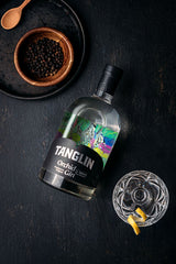 Tanglin gin bottle with a glass and bowl of black peppercorns on a dark surface