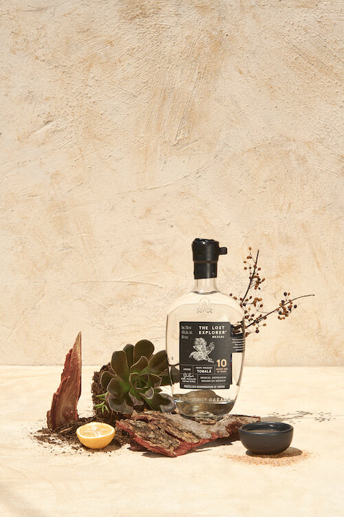 An artful display of The Lost Explorer Tobala Mezcal, set against elements of earth and rocks, a cacao bean, an agave plant, and a lemon half, with a textured pale background.