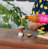Person pouring a yellow liquid from a bottle labeled 'Liber' into a metal cup on a wooden table.