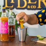 Person pouring a drink from a bottle labeled 'Liber & Co' into a metal shaker with bottles and limes in the background.