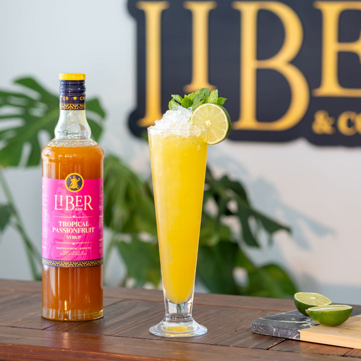 Bottle of Liber Tropical Passionfruit Syrup with a cocktail garnished with lime and whipped cream, set against a backdrop with 'Liber & Co' branding.