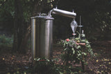 A moody image of a still and a vaporizor in the woods, a bottle of Trakal Patagonian Spirit on a rough-hewn stool next to the still, surrounded by herbs, leaves, and fruits.