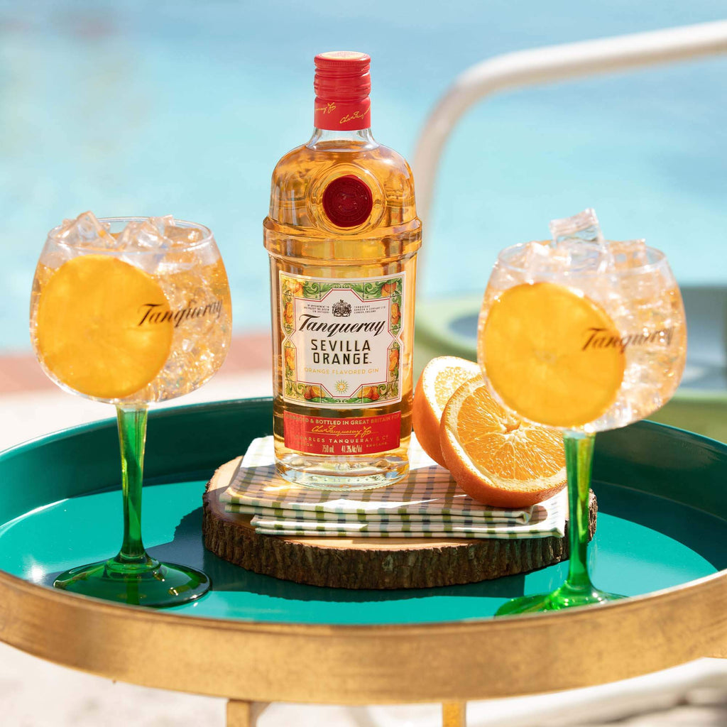 Two glasses of Tanqueray filled with ice and an orange wheel. They are flanking a bottle of Tanqueray Sevilla Orange on a teal colored platter, with fresh orange halves next to it.  From the background, the setting appears to be poolside.