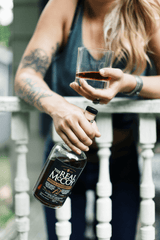A bottle of The Real McCoy 12 Year Rum, and a glass with one finger of rum poured inside it. Both being held by a woman wearing a casual gray tank top. She has blonde hair and her right arm is tattooed. She's leaning against a porch rail. The focal point of the image is the bottle and the glass with rum in it.