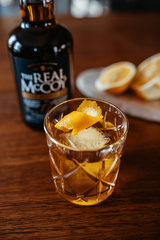A vintage glass holding amber liquid with an orange twist on top, likely an Old Fashioned. A bottle of The Real McCoy Single Blended Aged 12 Year in the background.