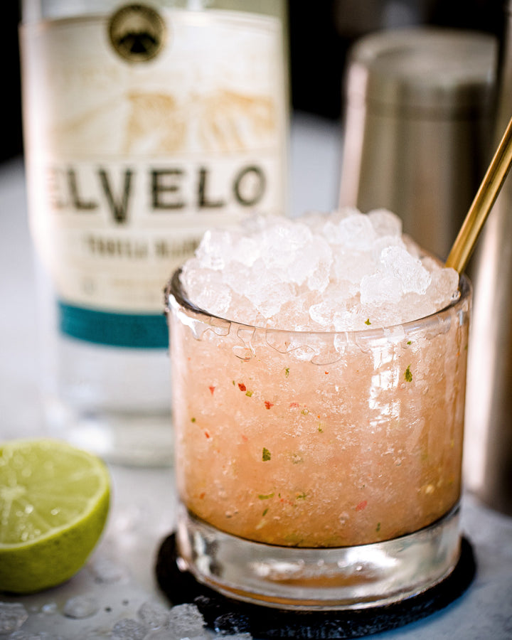 Glass of pink slushie with ice, bottle of Elvelo vodka, and lime on a table.