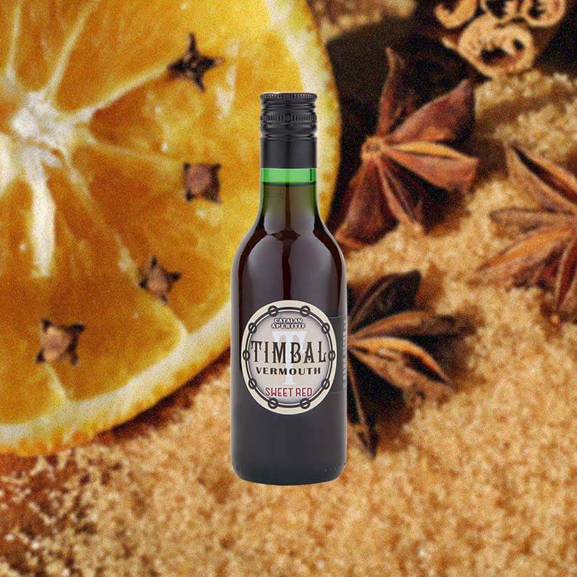 A bottle filled with deep ruby red liquid, reading Catalan Aperitif TIMBAL Vermouth Sweet Red on the label. It's a simple label and a smaller sized bottle - 500ml. Set again a macro shot of orange dotted with cloves, anise, cinnamon and rich brown sugar.