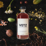 Bottle of VETZ Aperitivo Superiore over back drop image of assorted nuts, flowers, and spices on a dark table.