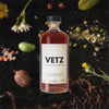Bottle of VETZ Aperitivo Superiore over back drop image of assorted nuts, flowers, and spices on a dark table.