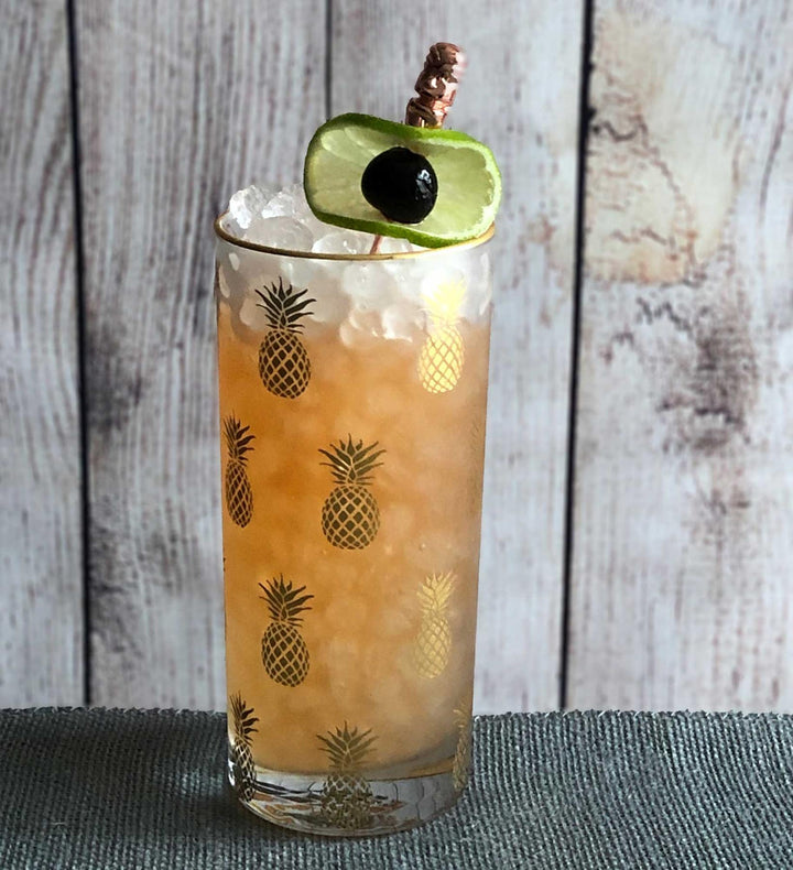 Cocktail in a glass with pineapple design, garnished with an avocado slice, on a wooden surface.