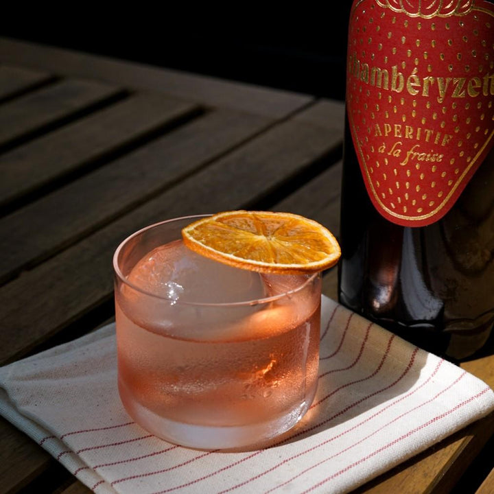 Glass of pink cocktail with an orange slice next to a bottle of Chamberyzet on a wooden table.