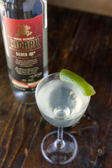 A bottle of Xicaru Silver 102 with a martini next to it.  Shot from slightly above, set on a wooden bar.