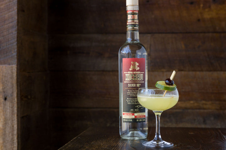 A bottle of Xicaru Silver 102 with a Last Word cocktail next to it.  On a wooden bar.