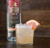 A bottle of Xicaru Silver 102 on a wooden bar, with a Paloma cocktail next to it.