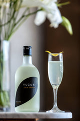 A bottle of Yuzuri with a tall floral arrangement in the background. Next to the bottle sits an elegant cocktail in a small flute, with a lemon twist on top.