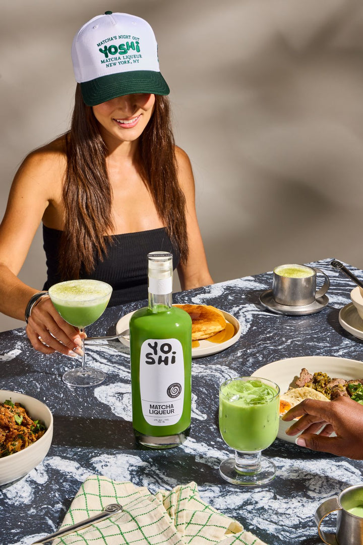 Woman holding a glass of green liquid at a table with food and a bottle labeled 'Nosh!'.