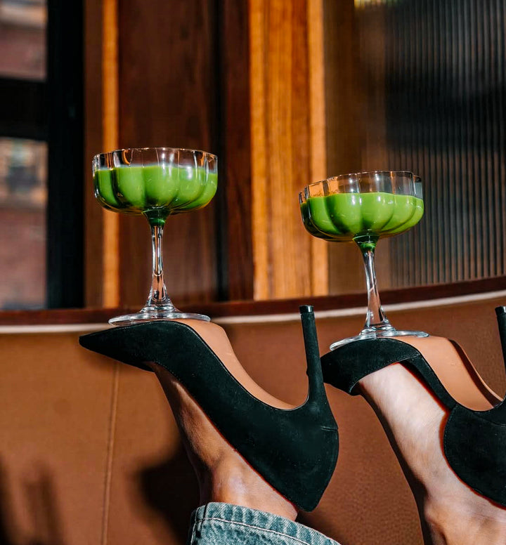 Green cocktails in coupe glasses held by black high heels on a blurred background