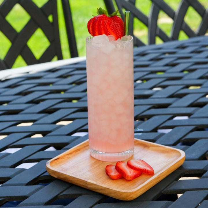 Pink cocktail with strawberries on a wooden coaster on a metal table outdoors.
