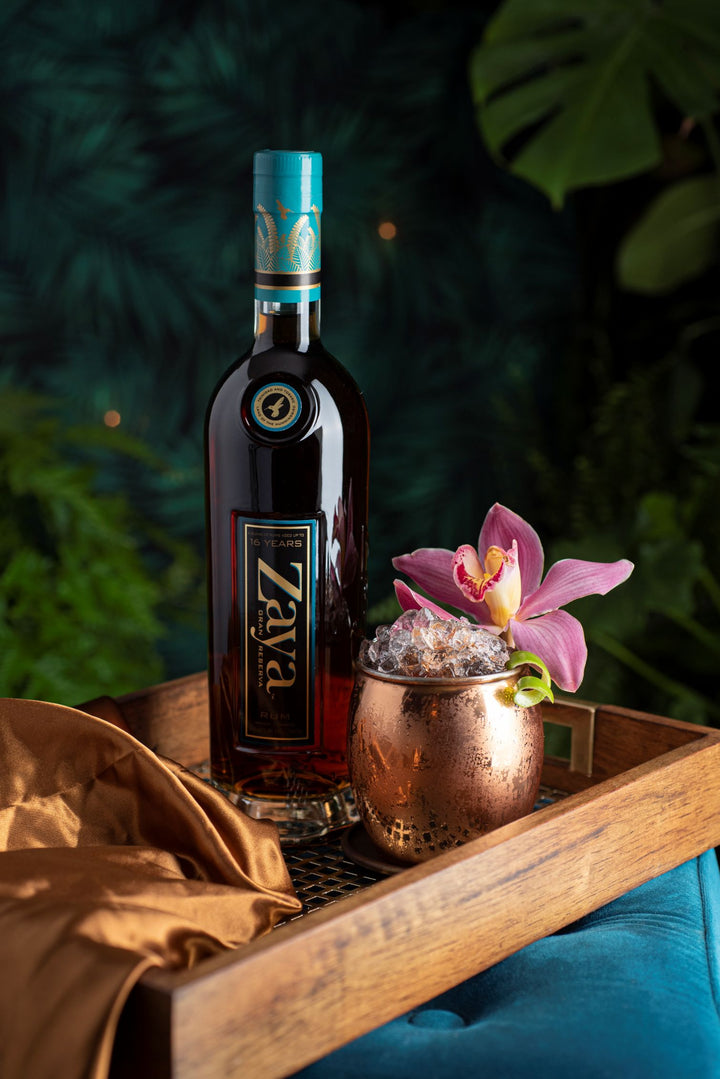 Bottle of Zaya rum with a copper mug and orchid on a wooden tray against a dark green leafy background