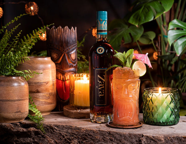 Zaya bottle with a cocktail garnished with mint and lime, surrounded by decorative items including candles and tiki masks.