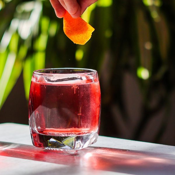 Red cocktail in a glass with an orange peel being squeezed over it, set against a blurred green outdoor background.