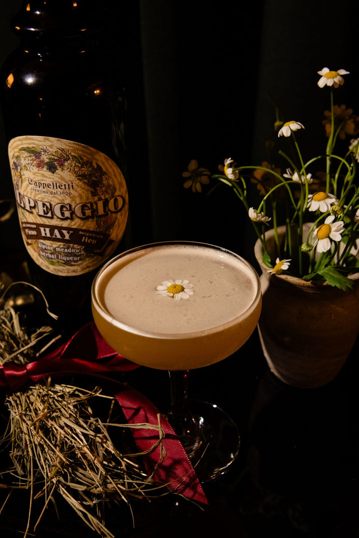 Cocktail with a flower garnish next to a bottle of Alpeggio Hay Liqueur and a vase of flowers on a dark background