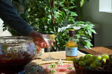 Person holding a glass of a drink with a bottle and grapes on a table outdoors.