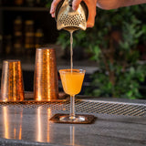Barista pouring a drink from a copper shaker into a glass on a bar.