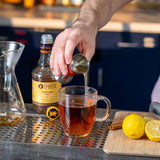 Person pouring a drink from a bottle into a glass on a bar with lemons and a cutting board in the background.
