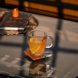 Clear glass mug with yellow liquid on a coaster on a reflective surface.