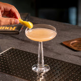 Cocktail in a coupe glass on a bar with a hand holding a yellow cocktail spoon.