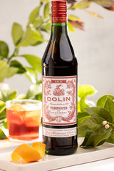 Bottle of Dolin Vermouth with a glass of cocktail and orange slices on a tray with green leaves in the background