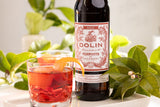 Bottle of Dolin Vermouth with a glass of red cocktail on a table with greenery.
