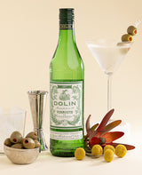 Green bottle of Dolin Vermouth with a cocktail glass and garnishes on a light background