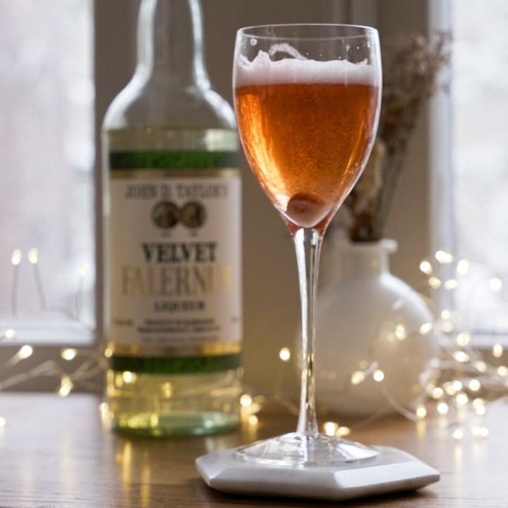 Glass of orange liquid with a bottle labeled 'Velvet Falernum' on a table with decorative lights.