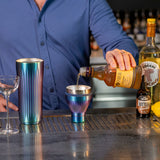 Bartender pouring a drink into a colorful metal shaker with bottles and glasses on a bar.