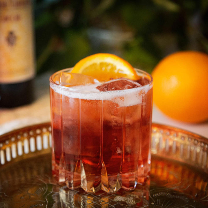 Red cocktail with an orange slice in a glass on a decorative tray with oranges and a bottle in the background.