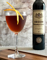 Cocktail in a glass with an orange twist next to a bottle of Cocchi Vermouth on a wooden surface.