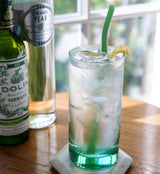 Glass of green cocktail with a lemon wedge and straw, next to bottles on a wooden table.