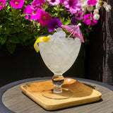 Glass of ice with a lemon wedge and pink umbrella on a wooden coaster, surrounded by flowers.