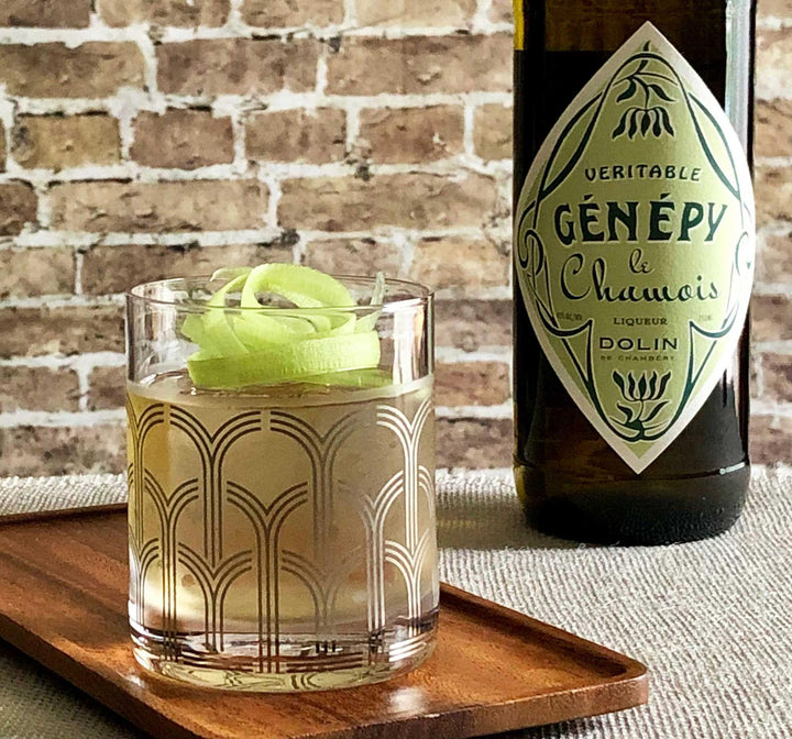 Glass of cocktail with a bottle of Genépy Le Chamois on a wooden tray against a brick wall.