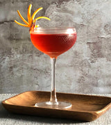 Red cocktail in a textured glass with an orange twist on a wooden tray against a gray stone background