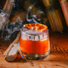 A macro shot of a cocktail with a deep orange color. One single large ice cube. A slice of smoked orange resting on top.  Fresh raw sugar cane stalks adorn the background.