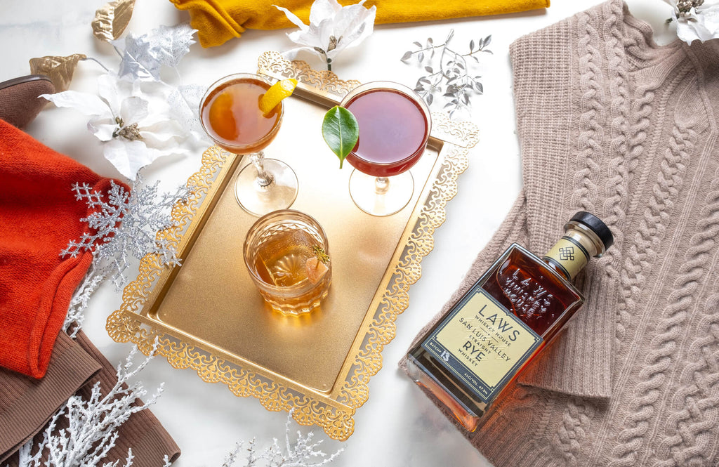 Bottle of Laws San Luis Valley Straight Rye on a table on top of a table cloth next to a tray with 2 cocktails.