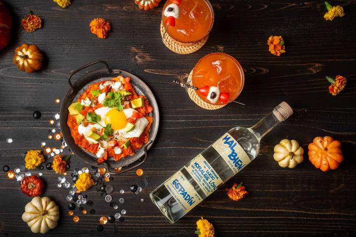 Bottle of Mal Bien Espadín Mezcal on a table laying label up next to 2 orange cocktails and delicious looking pan of food.