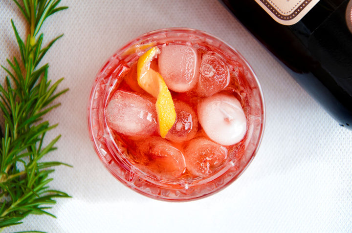 Cocktail with ice and lemon wedge on a white surface with a bottle and rosemary garnish.