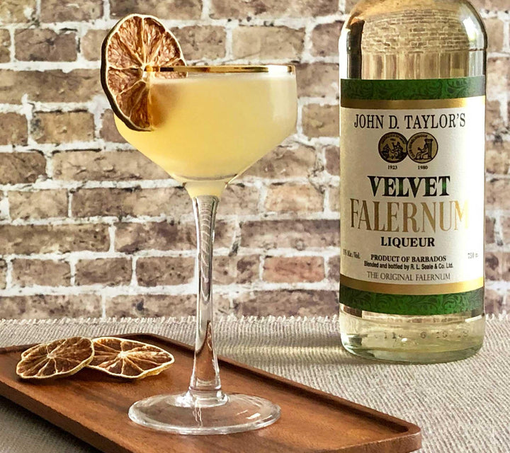 Cocktail with a lime wheel and bottle of John D. Taylor's Velvet Falernum Liqueur on a wooden tray against a brick wall.