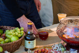 Person holding a cocktail next to a glass of red liquid on a table with grapes and other items.