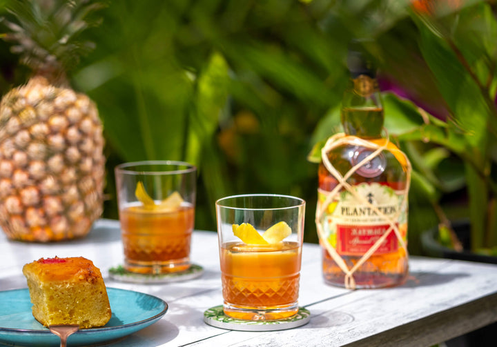 Bottle of Plantation Xaymaca Special Dry Jamaican Rum on a table outdoors next to 2 cocktails.