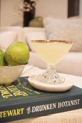 Cocktail glass with a drink on a book titled 'The Drunken Botanist' with limes in the background.